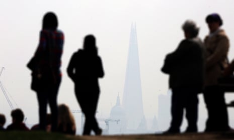People stand on Parliament Hill in Hampstead Heath overlooking an overcast London skyline.