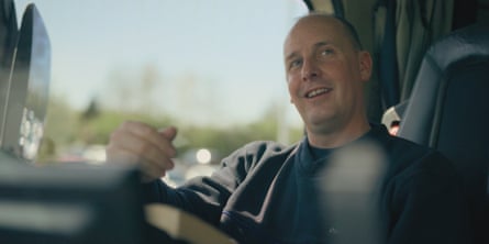 A close up of a smiling balding man wearing dark clothes sat in the driving seat of a lorry