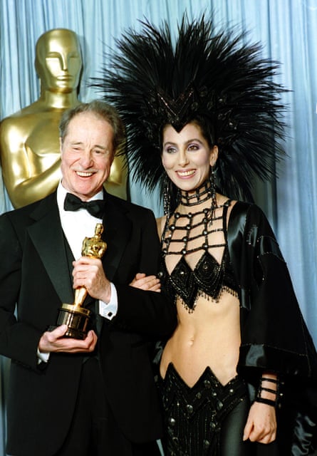 Cher wearing a black midriff-baring outfit with an oversized Mohawk headdress, standing next to actor Don Ameche