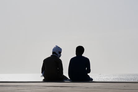 Two men, seen from behind in silhouette, sit on a beach wall.