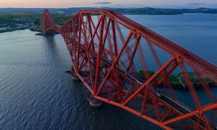 Aerial View of Train crossing the Forth Bridge at dusk, Queensferry, Edinburgh. This railway bridge, crossing the Forth estuary in Scotland, had the world’s longest spans (541 m) when it opened in 1890. It remains one of the greatest cantilever trussed bridges and continues to carry passengers and freight.