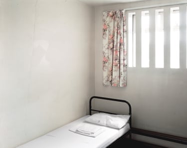 A prison cell with a single bed, folded sheets, and a floral curtain beside a barred window