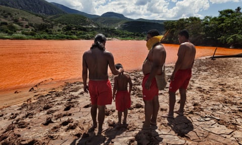 Three Indigenous men, one with a ruff of yellow feathers, stand with a child beside a wide river turned orange through pollution