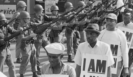 National Guard troops stand with bayonets fixed as African-American sanitation workers march wearing placards reading “I AM A MAN.”