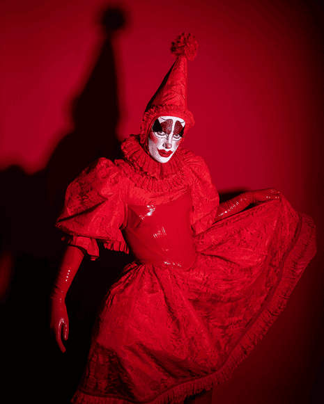 A person in all red - latex gloves, latex corset, dress with ruffled neck and puffed sleeves, and conical hat with poof on top, and red-and-white clown makeup.