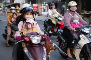 People on motorbikes in Hanoi. The adults are wearing helmets and some are wearing facemasks while the children sit in front and wear facemasks.