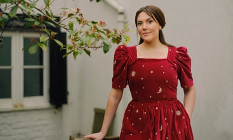 Katy Wix in a wine-red dress in a garden, cottage-like house behind
