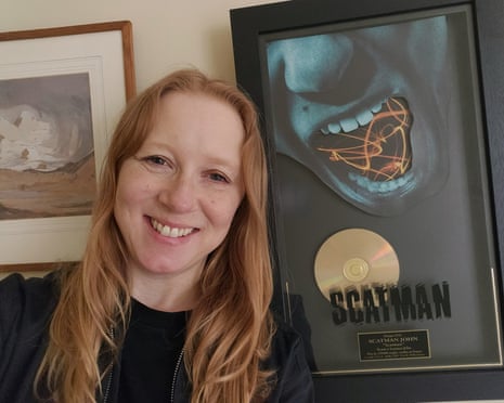A young woman with long red hair leaning into a poster showing a wide open mouth with gold wires inside and a gold CD