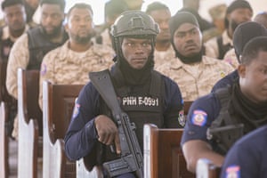 An armed police officer in a helmet and bulletproof vest sit on a pew surrounded by others in military fatigues