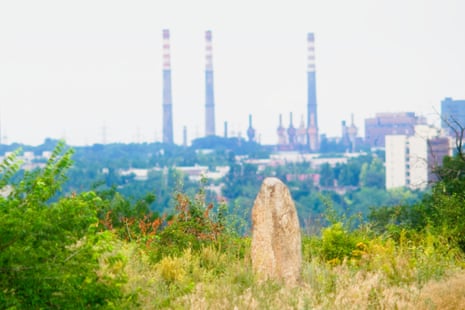 In the foreground is an obelisk surrounded by grass and bushes, while in the background are tall, dark smokestacks.