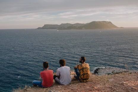 Three figures sitting by the sea looking out to an island.