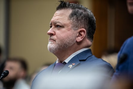 He sits in front of a microphone wearing a blue suit. Other people are pictured in the room, but out of focus