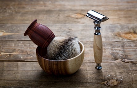 A shaving brush in a small dish and and a metal razor standing on end on a wooden table.