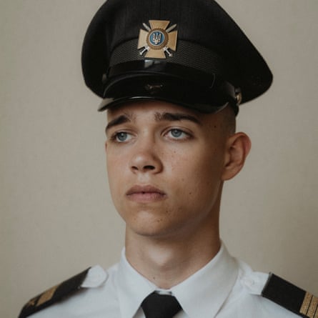 he has blue eyes and thick, dark eyebrows, and wears a tall cap with a metal cross-shaped badge and a white shirt with black epaulettes and a black tie. He looks serious and resolute.