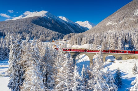Red train on a bridge amid the snowy landscape of Switzerland with mountains behind.