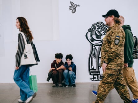 Two boys crouched on the floor looking at a phone screen as people walk past, including a man in military camouflage.