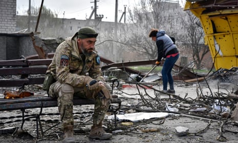 A Ukrainian serviceman smokes sitting on a bench as a local resident clears debris near a building damaged in the Russian air raid in the town of Orikhiv, Zaporizhzhia region, Ukraine, Friday, Apr. 5, 2024.