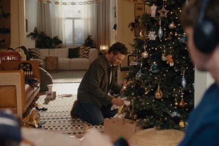 A man picks up a present from under a Christmas tree while a teenager with headphones is watching him