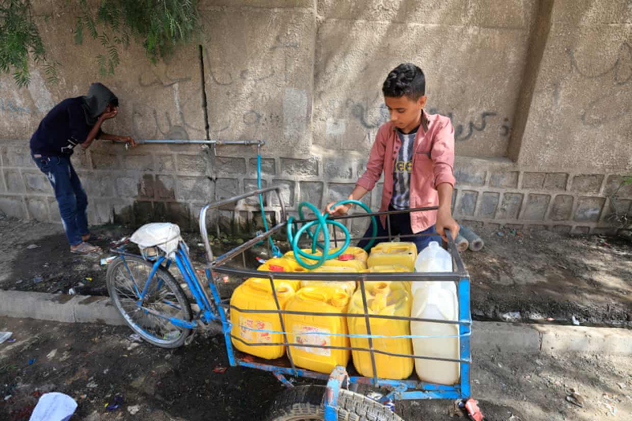 A boy fills plastic jerrycans with water as another drinks from a pipe. The cans are stacked in the front section of a cargo bicycle.