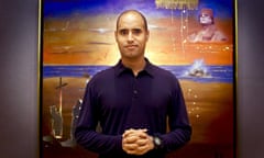 Saif al-Islam Gaddafi in 2002, standing in front of a painting that features an image of his father, Muammar Gaddafi, in the top right hand corner.