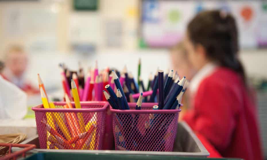 A typical UK primary school classroom with coloured pencils in the foreground and pupils in the background.