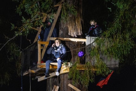 A man sits with his legs hanging off a structure of two-by-fours nailed into a treetop, with a spotlight shining on his from below.