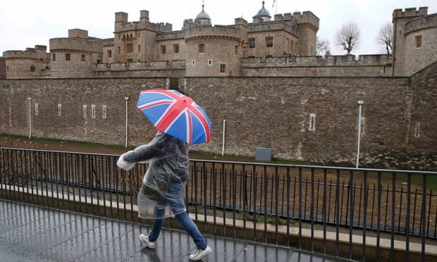 A man sheltering under a union jack umbrella walks past the Tower of London