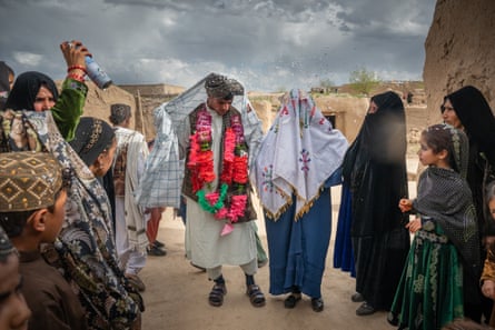 People surround a boy in a shalwar kameez with garlands of plastic flowers around his neck standing next to a girl with a large cloth over her head and shoulders. A woman sprays them with fake snow