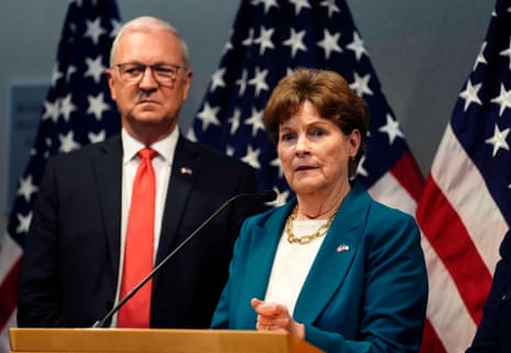 A woman with dark hair and a blue suit stands before American flags.