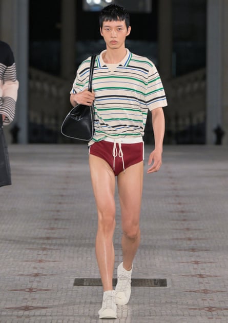 A model walks the catwalk wearing dark red Speedo-style swimming trunks, a striped T-shirt and white shoes. He carries a black holdall on his right shoulder.