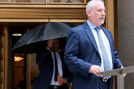 a man with grey hair and a grey beard in a blue suit and tie walks out of a gold doorway with a man behind him holding a dark umbrella