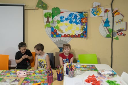 Two distracted boys and a girl aged about seven sit at a table covered with charts, pencils and pieces of paper, with children’s paintings on the wall behind them