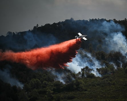 A plane flies low over a smouldering hillside dropping a thick cloud of red powder behind it.