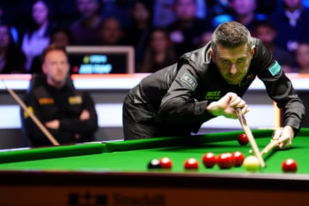 Mark Selby plays a shot while Judd Trump looks on from his seat.