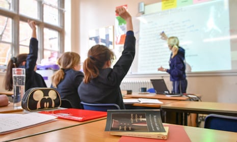 three schoolgirls, seen from behind, sit with raised hands in a classroom while an adult woman stands at a whiteboard