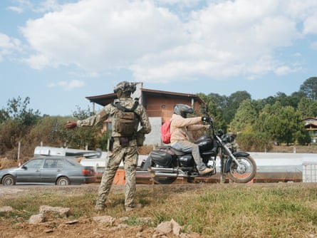 An army personnel guides traffic on a road as a motorcyclist rides past