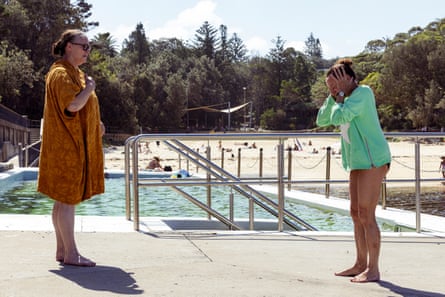 One woman in a tan dress and one woman in a mint-green top with her hands on her ears. They are standing by the steps of a swimming pool