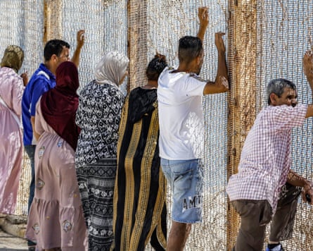 A row of people wait looking out through the wire-fence border at Ceuta