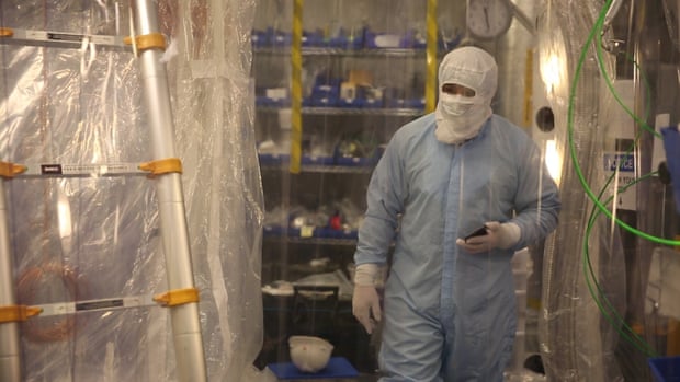 A lab worker wears a full body protective suit with head and face covering in a room covered in plastic sheeting.