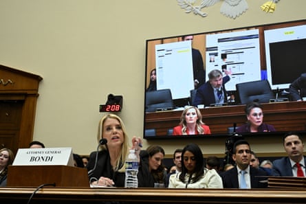 a woman speaking while seated at table, a TV screen behind her