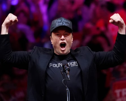 Elon Musk in a black Maga baseball cap, blue T-shirt and black suit jacket hollers into a microphone