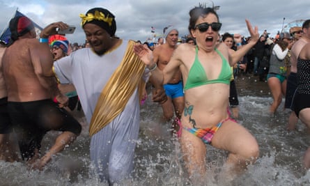 A Black man dressed in a white toga with gold leaf crown and gold sash, and a white woman wearing a bikini, appear to scream as they run into shallow water.
