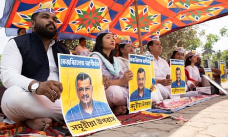 A row of people sitting cross-legged on the ground outside, holding posters showing Kejriwal’s face behind bars
