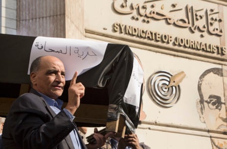 An Arab man gestures while holding a fake coffin draped in black and with placards attached to it. Behind him is a sign in Arabic and English saying 'Syndicate of Journalists'