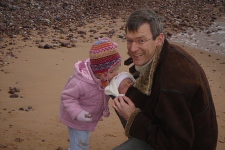 A man crouching down on a sandy and pebbly beach, with a baby in a sling and a little girl kissing the baby’s head
