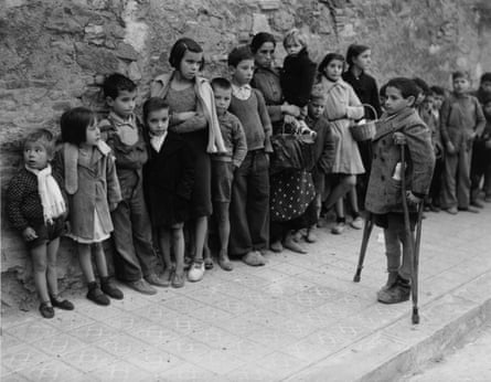 Children lean against a wall as the queue, with many of them looking at a boy on crutches who is stood separately on the pavement