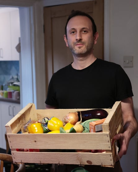 A man in a black T-shirt holds a crate of fruit of vegetables including yellow peppers and an aubergine