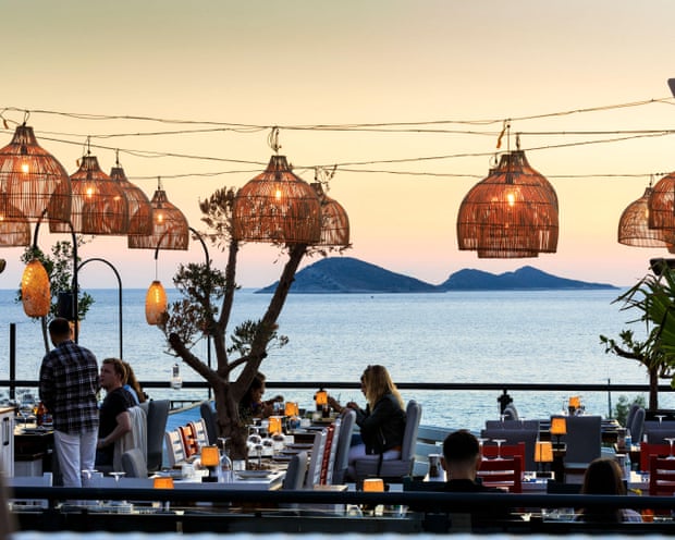 A seaside outdoor restaurant with hanging lamps and a view across the water to small islands