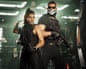 Jason and Lucia stand back-to-back holding rifles as cash flies through the air