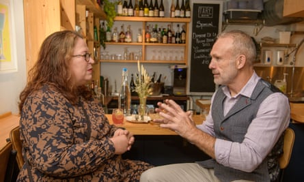 A woman with long red wavy hair, glasses and red lipstick, wearing a brown and black patterned dress, and a man with short grey hair, wearing a lilac shirt, a grey herringbone waistcoat and beige chinos, sitting at a restaurant table talking